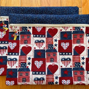 May include: Two quilted zipper pouches with a patriotic theme. The pouches feature a patchwork design with red, white, and blue squares, hearts, stars, and house motifs. The top edges are navy blue with white stars, and each pouch has a charm on the zipper.