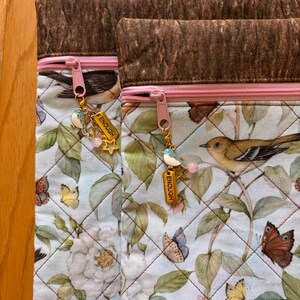 Puede incluir: Dos bolsas de tela acolchada con un estampado floral y de p&aacute;jaros. Las bolsas tienen cremalleras rosas y colgantes decorativos que dicen "ENOUGH". La parte superior de las bolsas es de tela marr&oacute;n oscuro texturizada.