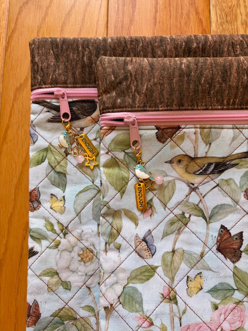 Puede incluir: Dos bolsas de tela acolchada con un estampado floral y de p&aacute;jaros. Las bolsas tienen cremalleras rosas y colgantes decorativos que dicen "ENOUGH". La parte superior de las bolsas es de tela marr&oacute;n oscuro texturizada.
