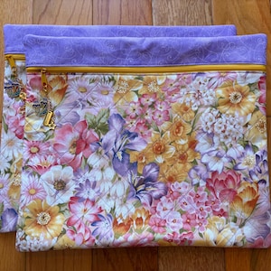 May include: Two quilted fabric pouches with a vibrant floral pattern in shades of pink, yellow, and purple. Each pouch has a lavender top with a gold zipper and decorative charms. The pouches are on a wooden surface.