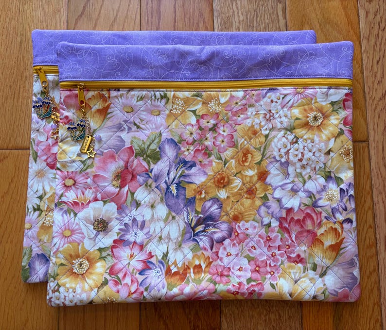 May include: Two quilted fabric pouches with a vibrant floral pattern in shades of pink, yellow, and purple. Each pouch has a lavender top with a gold zipper and decorative charms. The pouches are on a wooden surface.