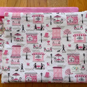 May include: A quilted fabric case with a pink and white cafe pattern. The design features pink cafe storefronts, tables, and people with dogs. A pink zipper and charm are visible on the left side. The case is on a wooden surface.