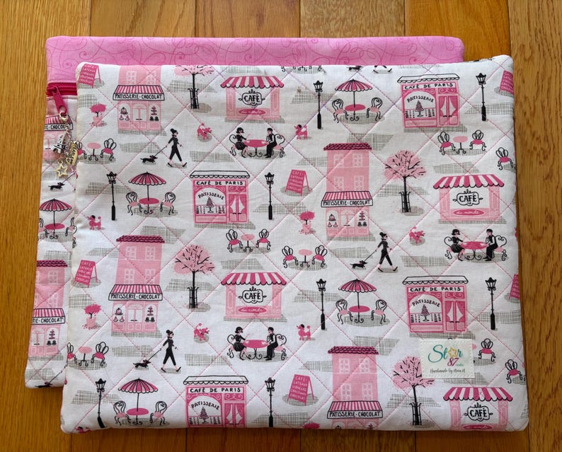 May include: A quilted fabric case with a pink and white cafe pattern. The design features pink cafe storefronts, tables, and people with dogs. A pink zipper and charm are visible on the left side. The case is on a wooden surface.