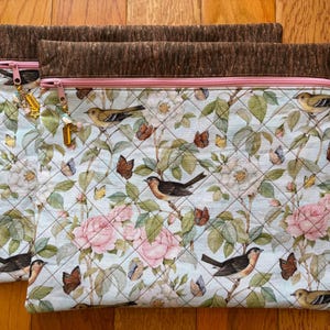 Puede incluir: Dos bolsas de tela acolchada con un estampado floral y de p&aacute;jaros. Las bolsas presentan un fondo azul claro con rosas rosas, mariposas y p&aacute;jaros. El borde superior es marr&oacute;n con una cremallera rosa y un dije. Sobre una superficie de madera.