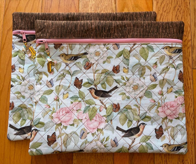 Puede incluir: Dos bolsas de tela acolchada con un estampado floral y de p&aacute;jaros. Las bolsas presentan un fondo azul claro con rosas rosas, mariposas y p&aacute;jaros. El borde superior es marr&oacute;n con una cremallera rosa y un dije. Sobre una superficie de madera.