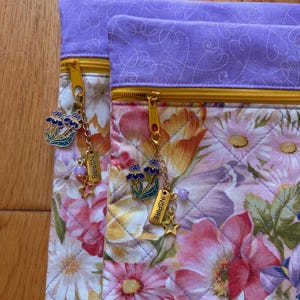 May include: Two floral print pouches with gold zippers and purple accents. Each pouch has a charm with a flower, star, and the word "INSPIRE". The pouches feature a quilted design with a variety of flowers in shades of pink, purple, and yellow.
