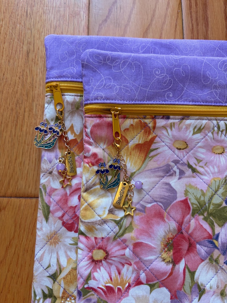May include: Two floral print pouches with gold zippers and purple accents. Each pouch has a charm with a flower, star, and the word "INSPIRE". The pouches feature a quilted design with a variety of flowers in shades of pink, purple, and yellow.
