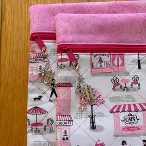 May include: Two pink and white quilted pouches with pink zips and charms. The fabric depicts a Parisian cafe scene with pink accents. Charms include an Eiffel Tower, stars, and the word "FEARLESS".