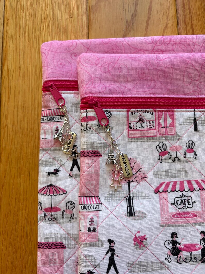 May include: Two pink and white quilted pouches with pink zips and charms. The fabric depicts a Parisian cafe scene with pink accents. Charms include an Eiffel Tower, stars, and the word "FEARLESS".