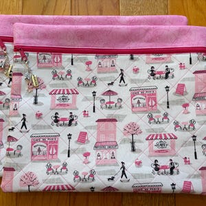 May include: Two quilted fabric pouches with a Parisian cafe print in pink, grey, and white. The pouches have pink zips and trim. The print features cafe scenes, people, dogs, and the Eiffel Tower. The pouches are on a wooden surface.