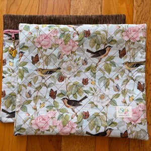 Puede incluir: Dos bolsas de tela acolchada. La bolsa superior presenta un fondo azul claro con un estampado floral de rosas rosas, p&aacute;jaros y mariposas. La bolsa inferior es de color marr&oacute;n liso. Una cremallera dorada con dijes es visible en la bolsa superior.