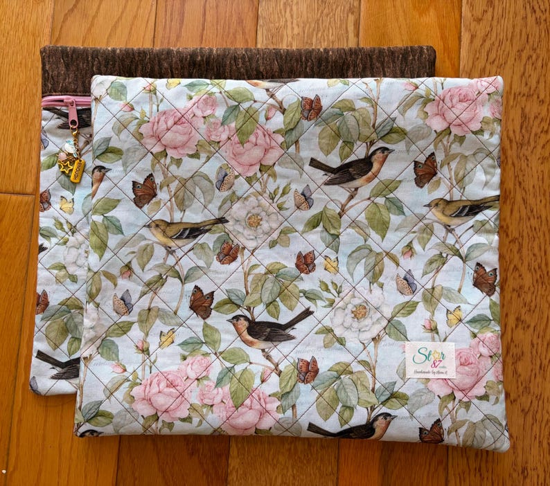 Puede incluir: Dos bolsas de tela acolchada. La bolsa superior presenta un fondo azul claro con un estampado floral de rosas rosas, p&aacute;jaros y mariposas. La bolsa inferior es de color marr&oacute;n liso. Una cremallera dorada con dijes es visible en la bolsa superior.