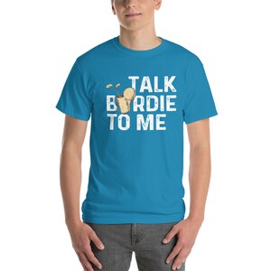 Op de afbeelding: Een turquoise blauw t-shirt met een witte afbeelding waarop staat "Talk Birdie To Me". De afbeelding toont een cartoon van een golfbal in een golfbeker.