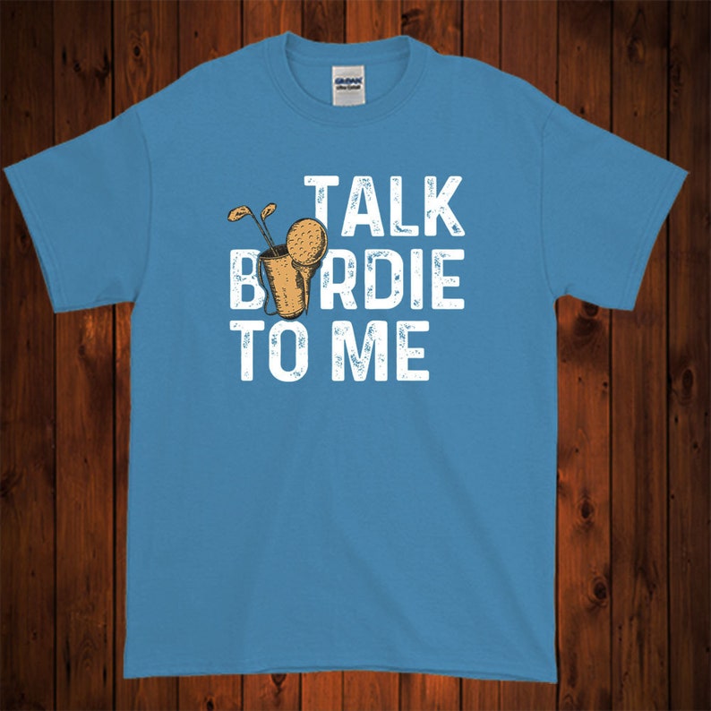 Op de afbeelding: Een lichtblauw t-shirt met een witte afbeelding waarop "Talk Birdie To Me" staat. De afbeelding bevat ook een cartoonafbeelding van een golfclub en een golfbal.