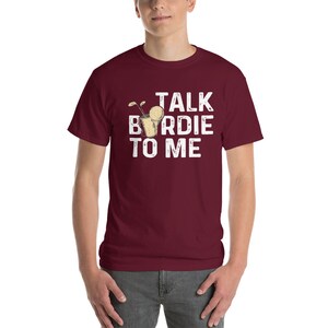 Op de afbeelding: Bordeauxkleurig T-shirt met korte mouwen met een witte afbeelding van een golfclub en een golfbal in een glas. De tekst "TALK BIRDIE TO ME" is in wit op het shirt gedrukt.