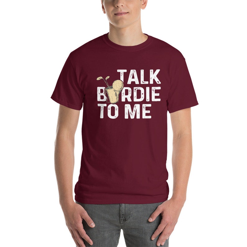 Op de afbeelding: Bordeauxkleurig T-shirt met korte mouwen met een witte afbeelding van een golfclub en een golfbal in een glas. De tekst "TALK BIRDIE TO ME" is in wit op het shirt gedrukt.