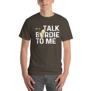 Op de afbeelding: Een bruin t-shirt met een witte afbeelding waarop "Talk Birdie To Me" staat. De afbeelding bevat een cartoon van een golfbal in een glas.