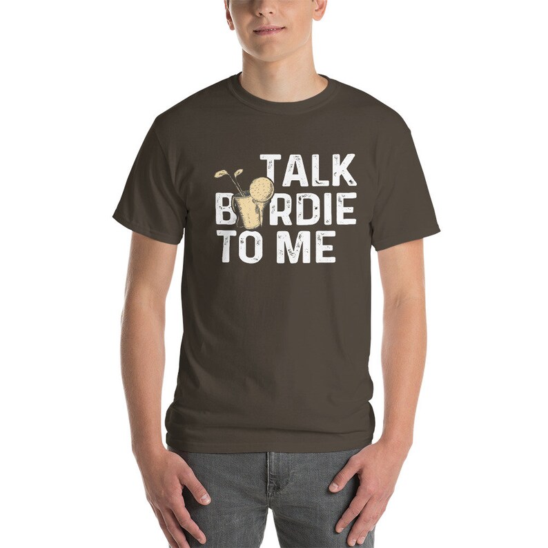 Op de afbeelding: Een bruin t-shirt met een witte afbeelding waarop "Talk Birdie To Me" staat. De afbeelding bevat een cartoon van een golfbal in een glas.