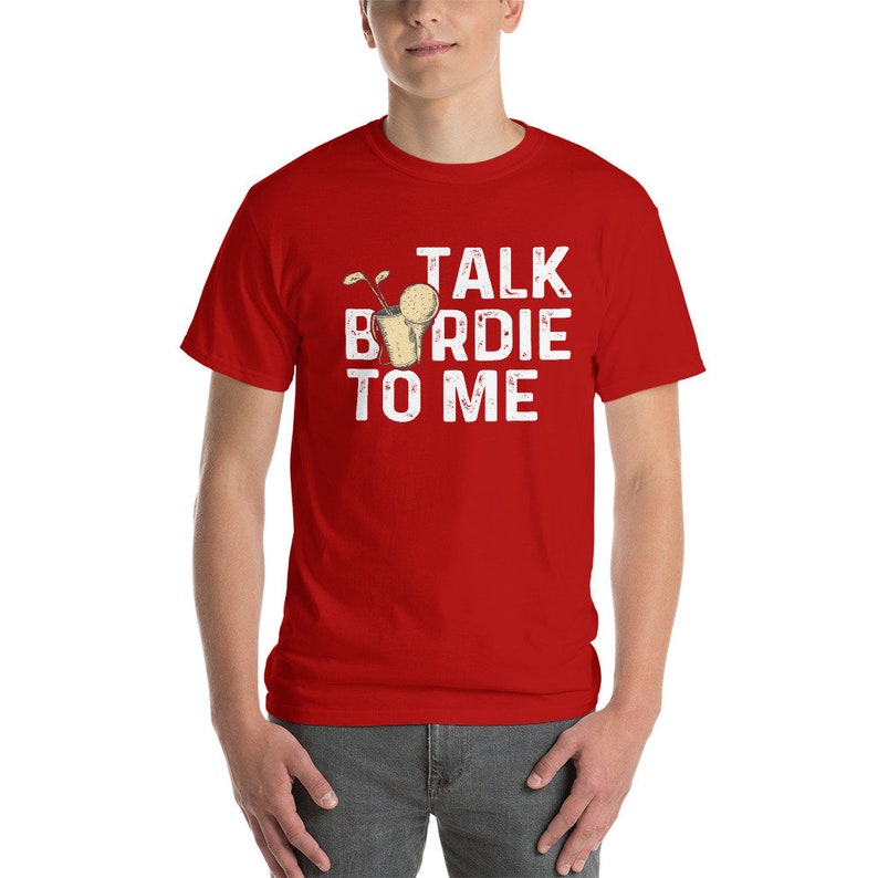 Op de afbeelding: Rood T-shirt met korte mouwen met witte tekst die "Talk Birdie To Me" luidt. Een golfbal met een steel zit in een glas.