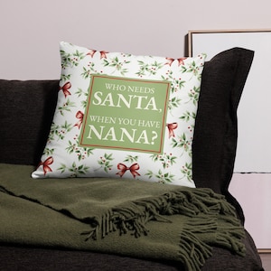 Puede incluir: Almohada blanca con un patrón de hojas de acebo verdes y rojas. La almohada tiene un rectángulo verde con el texto "¿Quién necesita a Santa, cuando tienes a Nana?"