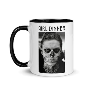 Peut inclure: Une tasse en céramique blanche avec un bord et une poignée noirs. La tasse présente une image en noir et blanc d'un homme avec un maquillage de crâne et le texte "GIRL DINNER" imprimé sur le devant.