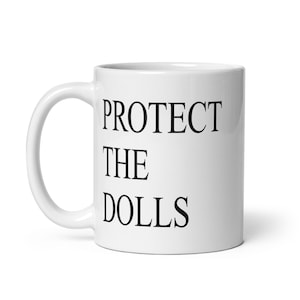May include: White ceramic mug with the text "PROTECT THE DOLLS" in black, bold letters. The mug has a curved handle and a cylindrical shape. The text is centered on the mug.