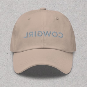 May include: A tan baseball cap with the word "COWGIRL" embroidered on the front in a light blue color.