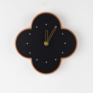 May include: A modern wall clock with a four-leaf clover shape. The clock face is black with white dot hour markers and gold-colored hands. The clock has a wooden frame.