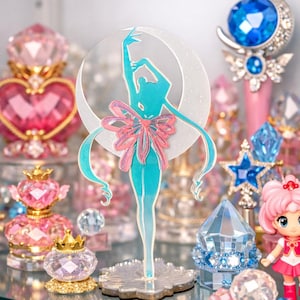 K&ouml;nnte beinhalten: T&uuml;rkisfarbene Silhouette einer Ballerina mit rosa Schleife, vor einem Halbmond. Umgeben von rosa und blauen Kristallfiguren, darunter ein Sternenstab und Parf&uuml;mflaschen, die eine skurrile Darstellung schaffen.