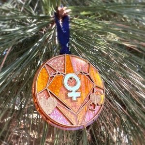May include: A round, wooden ornament with a stained glass design, featuring a light blue female symbol at the center. The ornament has orange, pink, and brown tones, with heart and floral accents. It hangs from a dark blue ribbon.