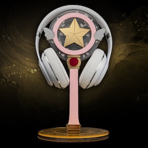 May include: White wireless headphones with silver detailing sit on a pink stand. The stand has a star-shaped design with a gold star. The base is gold and brown, set against a dark background.