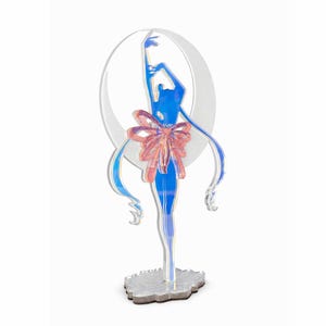 K&ouml;nnte beinhalten: Eine blau-rosa Acrylfigur einer Ballerina vor einem wei&szlig;en Halbmond. Die Ballerina hat lange, flie&szlig;ende Haare und eine gro&szlig;e rosa Schleife. Die Figur steht auf einem silbernen Sockel.