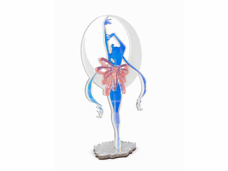 K&ouml;nnte beinhalten: Eine blau-rosa Acrylfigur einer Ballerina vor einem wei&szlig;en Halbmond. Die Ballerina hat lange, flie&szlig;ende Haare und eine gro&szlig;e rosa Schleife. Die Figur steht auf einem silbernen Sockel.