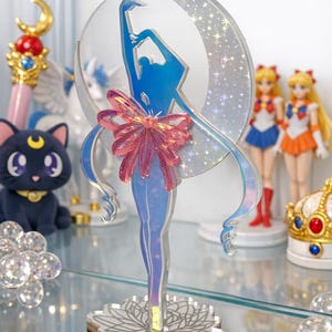 K&ouml;nnte beinhalten: Eine transparente Acrylfigur einer Ballerina in einer blauen Silhouette, mit einer rosa Schleife und einem mondf&ouml;rmigen Hintergrund. Die Figur steht auf einem floralen Sockel, mit anderen Anime-Themen im Hintergrund, darunter Puppen und eine Katze.