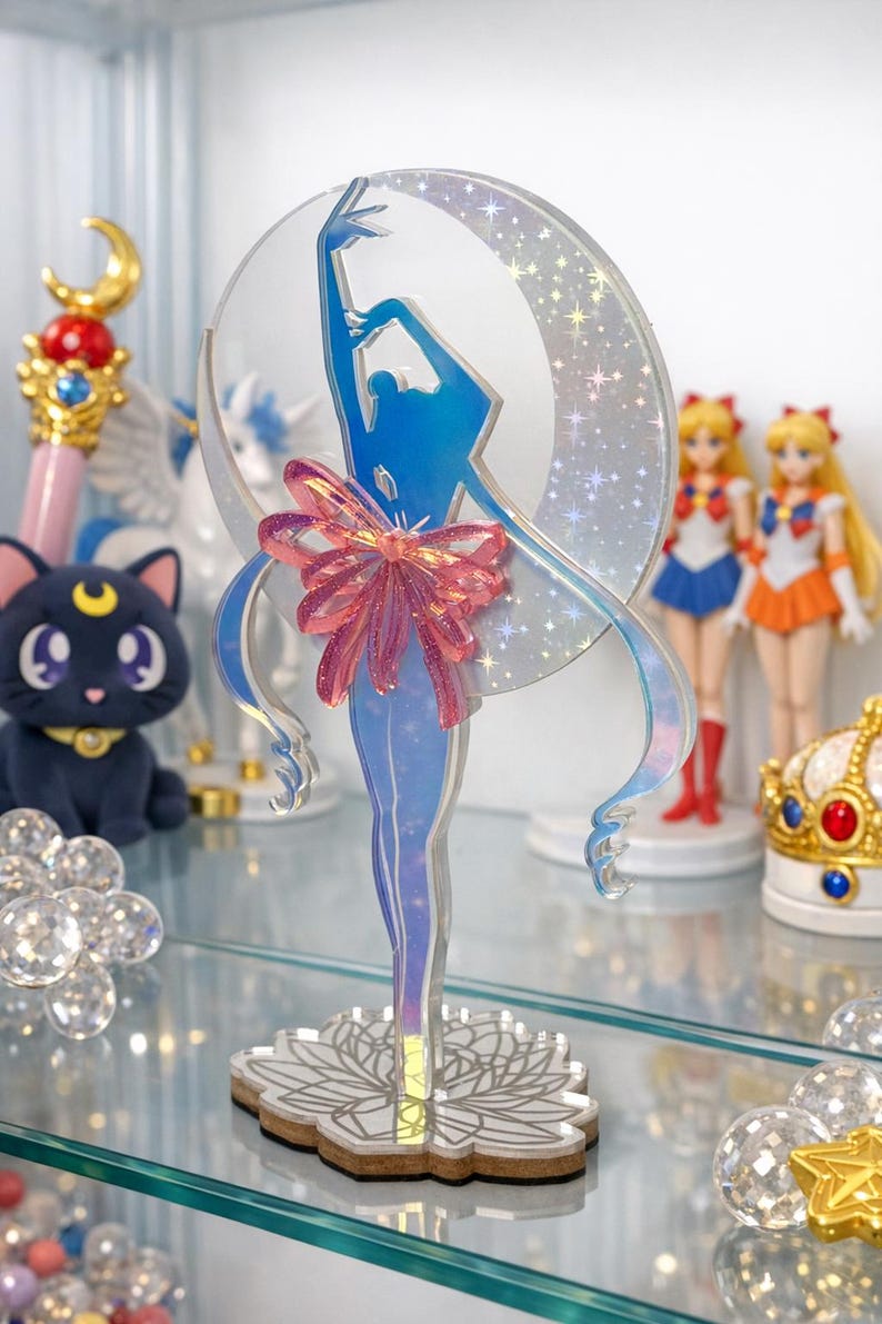 K&ouml;nnte beinhalten: Eine transparente Acrylfigur einer Ballerina in einer blauen Silhouette, mit einer rosa Schleife und einem mondf&ouml;rmigen Hintergrund. Die Figur steht auf einem floralen Sockel, mit anderen Anime-Themen im Hintergrund, darunter Puppen und eine Katze.