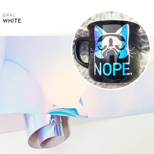 May include: A black ceramic mug with a white cat graphic and the word "NOPE" in blue iridescent vinyl. The mug is sitting on a white surface with a roll of iridescent vinyl in the background.