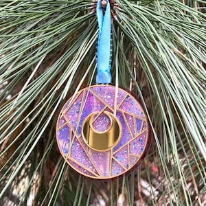 May include: A round Christmas ornament with a geometric design in shades of purple, pink, and gold. The ornament is hanging from a blue ribbon against a backdrop of green pine needles. The ornament has a gold crescent moon shape.
