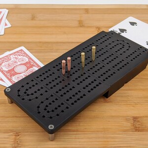 May include: A black cribbage board with metal pegs is displayed on a wooden surface. Playing cards are visible in the background. The board has a series of holes for scoring and is designed for the card game cribbage.