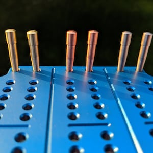 Puede incluir: Seis varillas metálicas con extremos redondeados se insertan en una placa de metal azul perforada. Las varillas son de diferentes tonos de cobre y oro.