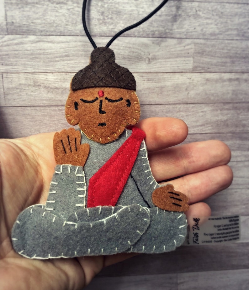 Op de afbeelding: Een vilten Boeddha-hanger in een zittende meditatiehouding. De hanger heeft een bruin gezicht met gesloten ogen, een rode stip en een donkerbruine knot. De Boeddha draagt een grijze jurk met een rode sjerp en is gestikt met witte draad. Een zwart koord is bevestigd.