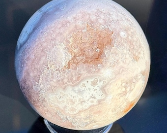 May include: A pink and white agate sphere with a natural crystal cave opening. The sphere is on a clear glass stand.