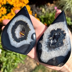 Puede incluir: Dos rodajas de ágata pulida en forma de lágrima con bordes exteriores de color negro intenso y bandas concéntricas de estructuras cristalinas blancas y grises que rodean centros huecos y brillantes con formaciones minerales.