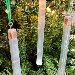 May include: Three selenite crystal ornaments hanging on a Christmas tree. Each crystal is a long, clear, rectangular shape with copper wire wrapped around the top. Green ribbon is used for hanging.