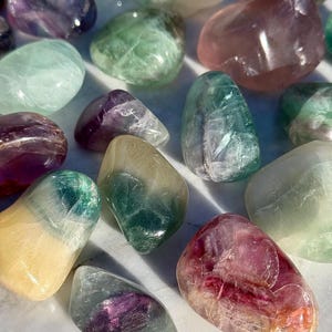 May include: An assortment of polished fluorite stones in various colors, including green, purple, pink, and yellow. The stones have a smooth, rounded shape and a translucent appearance, with visible internal patterns. The arrangement is scattered, showcasing the natural beauty of the crystals.
