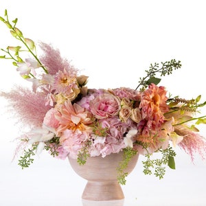 May include: A pink and peach floral arrangement in a light brown ceramic vase. The arrangement includes roses, hydrangeas, dahlias, and pampas grass.