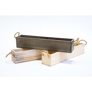 May include: Three wooden planter boxes with rope handles. The boxes are stacked on top of each other. The top box is dark brown, the middle box is light brown, and the bottom box is natural wood.