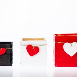 May include: Three wooden boxes with different colored paint finishes. Each box has a heart-shaped decoration hanging from a string. The boxes are black, white, and red.