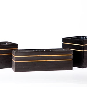 May include: Three black wooden planter boxes with gold trim. The boxes are different sizes and are arranged in a row on a white surface.