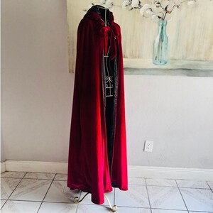 May include: A floor-length, deep red velvet cloak featuring a hood and a tie closure at the neck. The garment is lined with a contrasting black fabric and is displayed on a wire mannequin stand, showcasing its full length and draped silhouette.