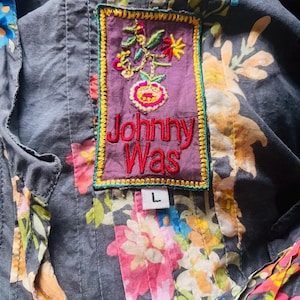 Puede incluir: Primer plano de una camisa con estampado floral y un parche decorativo. El parche es morado con flores bordadas y el texto "Johnny Was" en rojo. La camisa es gris con un estampado floral colorido y una etiqueta de talla "L".